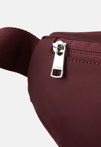 Borsa in nylon borgogna con superficie testurizzata, dotata di una cerniera argentata e branding. Design compatto con una tracolla in tessuto spesso per il fissaggio.