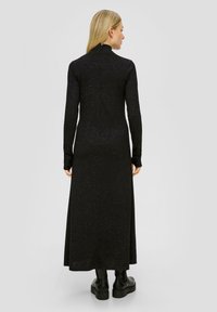 Robe noire à manches longues avec un col montant et une fermeture éclair dans le dos. Tissu doux et texturé avec de subtils éclats. Portée avec des bottes noires arrivant aux chevilles.