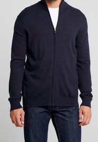Marinblå zip-up cardigan gjord av mjukt stickat tyg, med ståkrage, ribbade manschetter och nederkant, som bärs över en vit t-shirt.