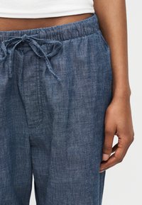 Tuvplāns uz cilvēku, kas valkā zilas lina bikses ar auklu un baltu cropped topu, roka atrodas blakus.