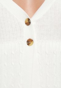 Bílý pletený kardigan s vzorem copánků, s dvěma kulatými tortoise-shell knoflíky na výstřihu pro zapínání.