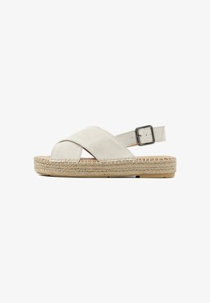 Beige suède sandalen met kruislings gespen, een gewaxt jute zool en een verstelbare metalen gesp op de enkelriem.