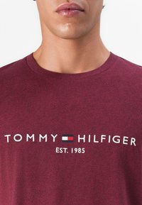 Zbliżenie na osobę w bordowej koszulce z logo "Tommy Hilfiger" i napisem "Est. 1985" na piersi.