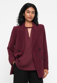 Blazer bordeaux de dupla abotoadura com botões pretos, lapelas afiadas e uma textura suave, combinado com uma blusa bordeaux a condizer.