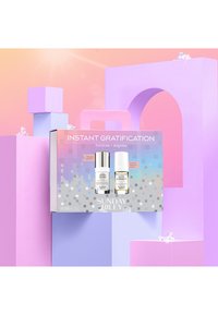 Ensemble de deux produits de soins de la peau dans une boîte argentée et grise, avec une bouteille blanche et une bouteille accentuée d'or sur un fond géométrique coloré.