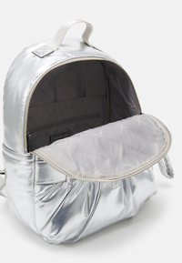 Mochila metálica plateada con forma redondeada, forro interior acolchado, bolsillo frontal y cierre de cremallera en contraste.