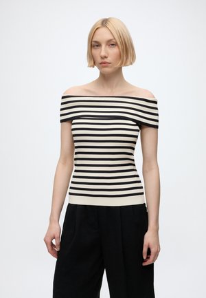 Femme aux cheveux blonds courts portant un haut à rayures horizontales noir et blanc, épaules dénudées, et un pantalon noir, debout devant un fond uni.
