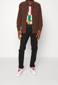 Braunes Cordhemd mit zwei Brusttaschen, getragen über einem mehrfarbigen, gemusterten Pullover, kombiniert mit schwarzer Hose und weißen Sneakers mit roten Akzenten.