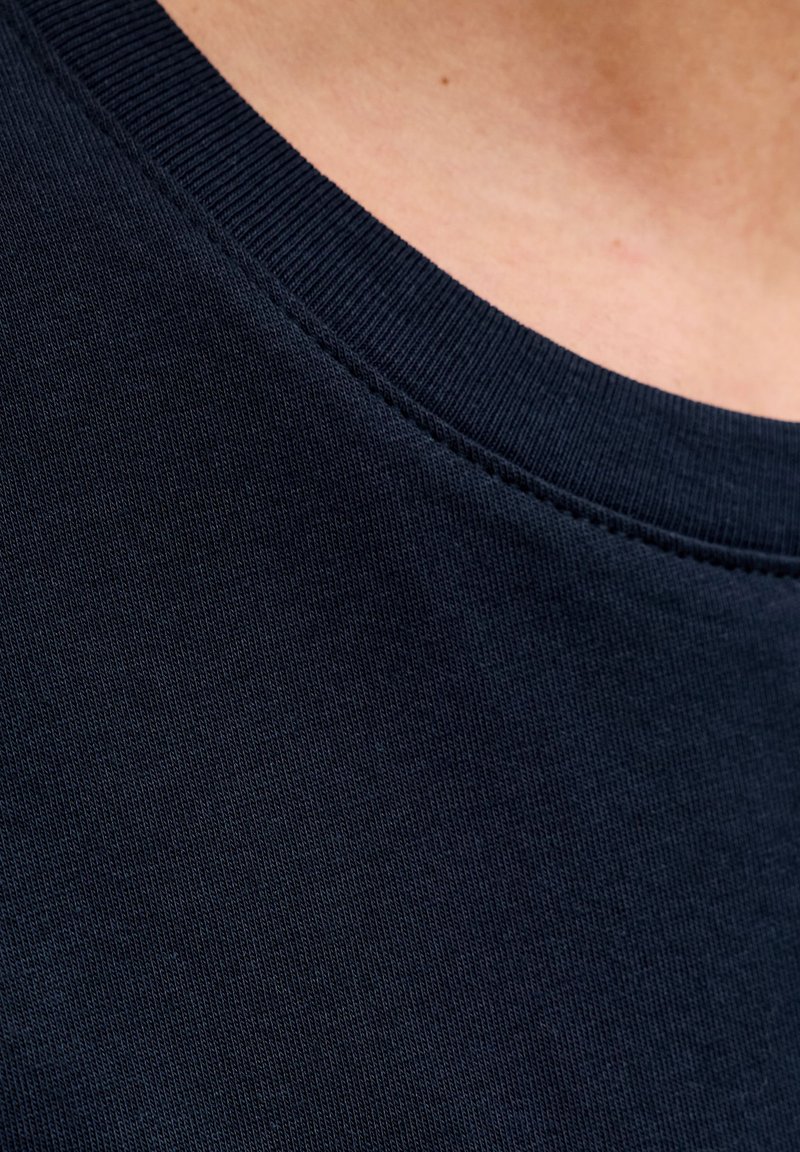 Mornarsko modro bombažno blago z gladko teksturo, s krožnim izrezom in rebrastim okrasjem ovratnika. Šivi so vidni vzdolž robov.