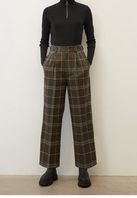 Svart zip-neck långärmad topp i kombination med vida höga byxor med rutmönster i bruna, gula och blåa ränder; svarta ankelboots.