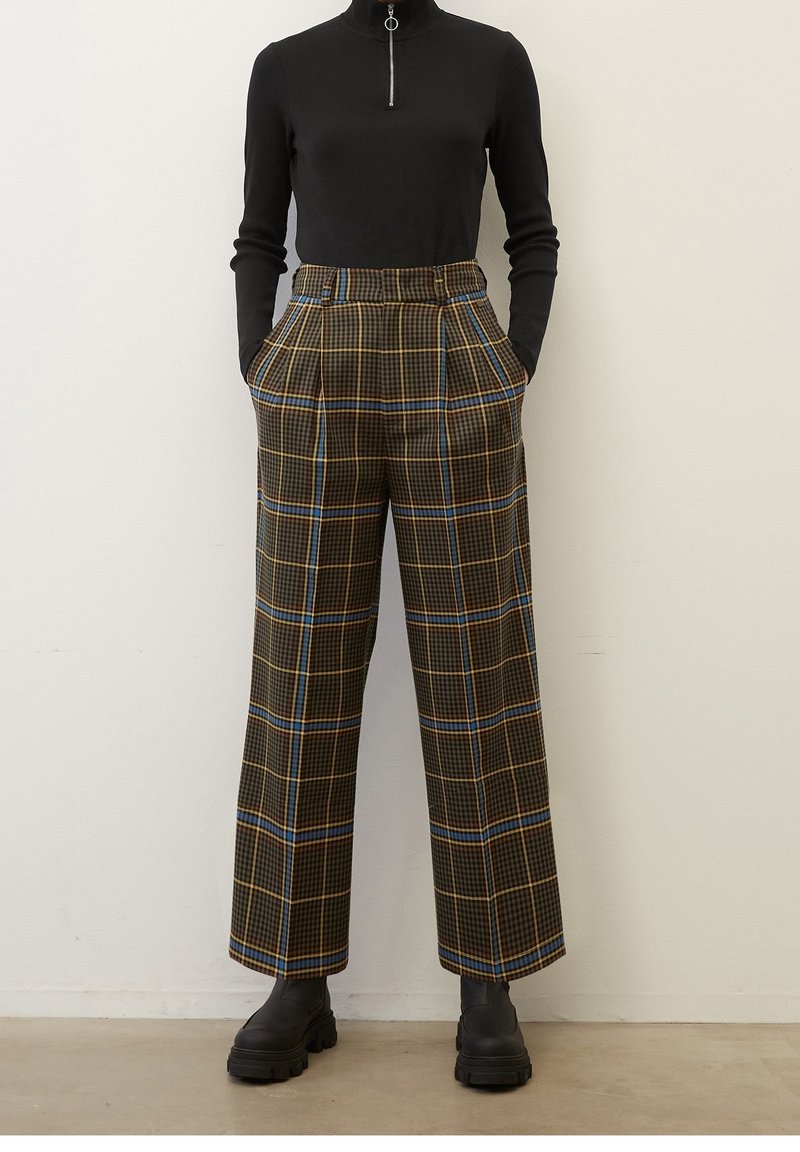 Svart zip-neck långärmad topp i kombination med vida höga byxor med rutmönster i bruna, gula och blåa ränder; svarta ankelboots.