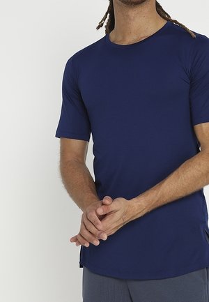 Mężczyzna o jasnobrązowej skórze i dredach, ubrany w przylegający granatowy t-shirt z krótkim rękawem i szare spodnie, z rękami splecionymi na przodzie.