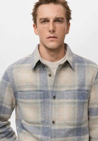 Karirana flanel košulja u bež i svijetlo plavoj boji, s dugmadi sprijeda, šiljastim ovratnikom i dva džepa na prsima s tamnim dugmadi.