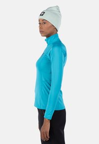  Lichtblauw hardloopshirt met lange mouwen en ritskraag, aansluitend ontwerp en subtiele logo op de borst; gecombineerd met zwarte broek en een lichtkleurige beanie.