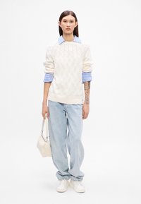 Krémový sveter s pleteným vzorom, ktorý je vrstvený cez svetlomodrú pruhovanú košeľu, v kombinácii so svetlomodrými voľnými džínsami a bielymi teniskami. Drží malú bielu tašku.