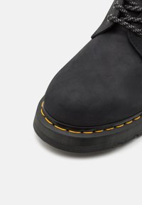 Botine negre din suede, cu suprafață texturată, vârf rotund și talpă din cauciuc cu cusături galbene. Șireturile sunt de culoare închisă și au un model cu stropi.