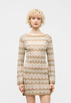 Kvinde med kort krøllet blondt hår iført en tætsiddende langærmet kjole med chevronmønster i beige, brun og pastel farver mod en hvid baggrund.