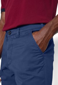 Mörkblå chinos av bomull med knappstängning och sidofickor, ihop med en vinröd polo-tröja. Synlig hand i fickan.