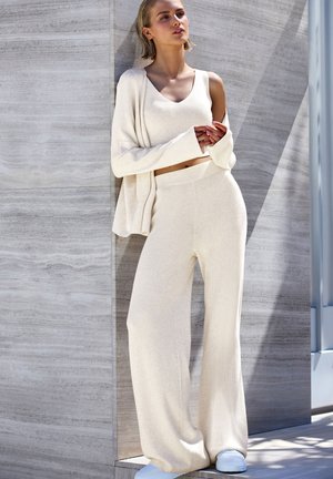 Hellbeige gerippter Strick-Loungewear-Set bestehend aus einem Oberteil mit Rundhalsausschnitt und weit geschnittenen Hosen, kombiniert mit weißen Sneakers vor einem steinernen Hintergrund.