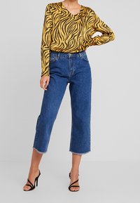 Donna che indossa un top a maniche lunghe giallo e nero con stampa zebra, jeans cropped blu a vita alta con orli sfrangiati e tacchi alti neri con cinghie.