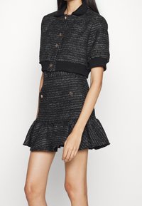 Svart tweedkort jacka med krage och knappstängning, kombinerad med en matchande kjol med volangkant och dekorativa knappar.