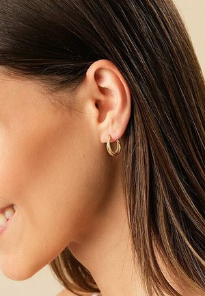 Goldene Creolen mit einem gedrehten Design, die eine glatte metallische Textur aufweisen, präsentiert am Ohrläppchen eines Models vor einem neutralen Hintergrund.