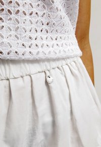 Weißes Baumwollhemd mit rautenförmigen durchbrochenen Mustern und einem gerafften elastischen Bund in einem weißen Rock, der einen kleinen silbernen Anhänger trägt.