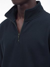 Homme portant un pull marine foncé à fermeture éclair quart de zip avec le col partiellement descendu, montrant un gros plan du cou et du haut de la poitrine.