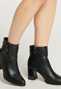 Botine negre din piele, cu tocuri groase, fermoare laterale și detalii decorative din scoică țestoasă pe cataramă și toc, purtate pe picioare goale.