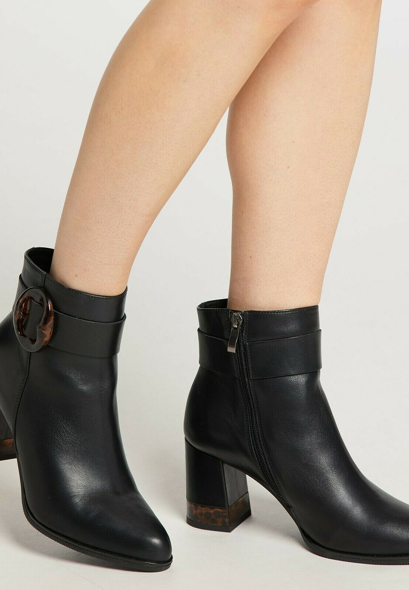 Botine negre din piele, cu tocuri groase, fermoare laterale și detalii decorative din scoică țestoasă pe cataramă și toc, purtate pe picioare goale.