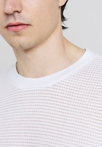Weißer strukturierter Pullover mit rundem weißen Kragen, der ein waffelartiges Strickmuster aufweist. Detail an dem glatten Ausschnitt und dezente Nähte.