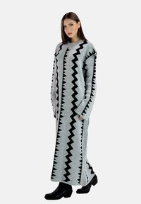 Lang grå strikket kjole med sort zigzagmønster, brede ærmer og afslappet pasform, kombineret med sorte ankelstøvler.