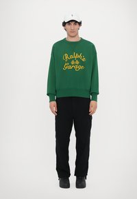 Green sweatshirt with yellow "Ralph's Garage" print, ribbed cuffs, and hem; paired with black trousers and black shoes; cap with logo.
