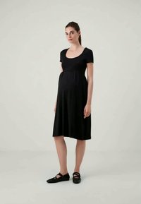 Melnā grūtniecības kleita, izgatavota no mīkstas auduma, ar izgriezumu uz dekoltē, īsām piedurknēm un pieguļošu impērijas vidukli, kas plūst uz ceļgala garuma svārkiem.