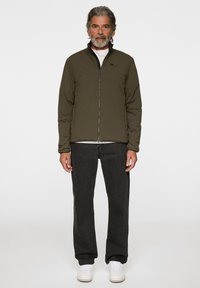 Olivgrön lättviktsjacka med ståkrage, dragkedja och subtil logotyp på bröstet, matchad med svarta jeans och vita sneakers.