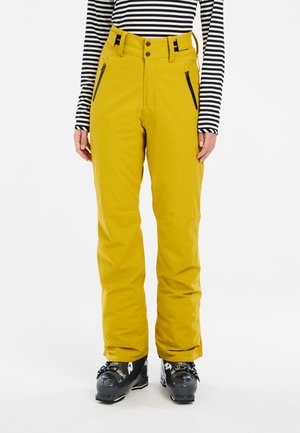 Gelbe Schneehosen mit hoher Taille, verstellbaren Trägern und seitlichen Reißverschlusstaschen, kombiniert mit schwarzen Skischuhen.