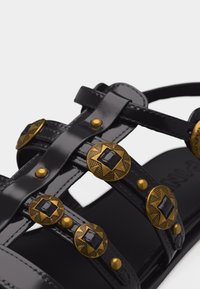 Lanières de sandales noires brillantes ornées de clous ronds en métal doré avec des motifs d'étoiles et des embellissements centraux noirs.