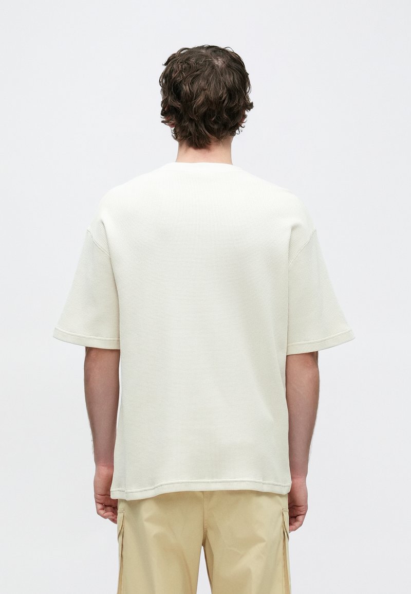 White, textured short-sleeve t-shirt with a crew neck, visible stitching, and relaxed fit, paired with light beige pants.