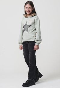 Sudadera verde claro con un diseño de estrella y capucha de estampado de leopardo, combinada con jeans negros que tienen tachuelas doradas a lo largo de las costuras laterales y botas negras.
