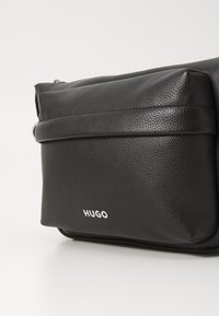 Borsa a tracolla in pelle nera texture con una tasca frontale con zip e logo bianco "HUGO" al centro della tasca.