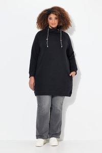 Donna con capelli ricci che indossa una felpa nera con cappuccio a maniche lunghe, jeans grigi e scarpe da ginnastica bianche, in piedi davanti a uno sfondo bianco.