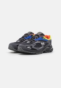Pari harmaita New Balance -urheilujalkineita mustilla pohjilla, sinisillä ja keltaisilla yksityiskohdilla sekä oranssilla sisävuorella valkoista taustaa vasten.