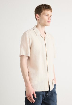 Short-sleeve beige linen shirt with a relaxed fit, a notched collar, and front buttons, paired with dark denim jeans.
