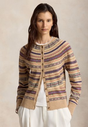 Femme aux longs cheveux foncés portant un cardigan beige à motifs boutonné sur une chemise blanche et un pantalon blanc, debout devant un fond neutre.