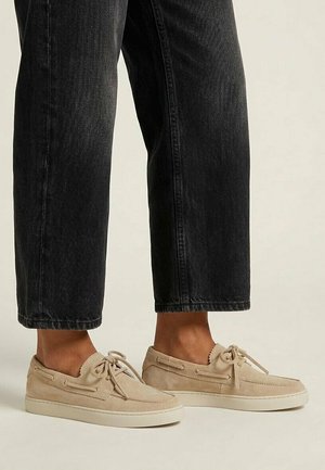 Beige suede moccasin shoes with laces worn with black cropped jeans on a light background.