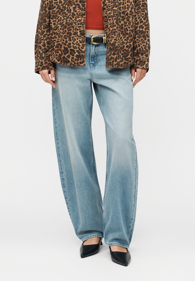 Άτομο που φοράει ανοιχτό μπλε τζιν παντελόνι με φαρδιές γραμμές, μαύρες μυτερές γόβες, ένα τζάκετ με λεοπάρ μοτίβο και ένα κόκκινο κοντό τοπ με μαύρη ζώνη.