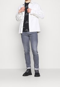 Vit denimjacka över en svart t-shirt med vit text, tillsammans med ljusgrå jeans och svarta sneakers. Modell som står i en neutral miljö.