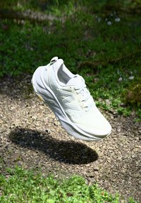 Witte sportieve schoen met een getextureerde mesh bovenzijde, gevoerde rand en vetersluiting, met een gestoffeerde zool en logo-accent aan de zijkant.