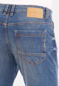 Vaqueros de mezclilla en azul medio, con un parche de cuero en la cintura, costuras de color cobre y un único bolsillo trasero con un detalle de marca.