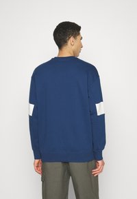 Blue sweatshirt with long sleeves, round neck, and white stripe details on sleeves. Smooth texture, casual fit, and ribbed cuffs.